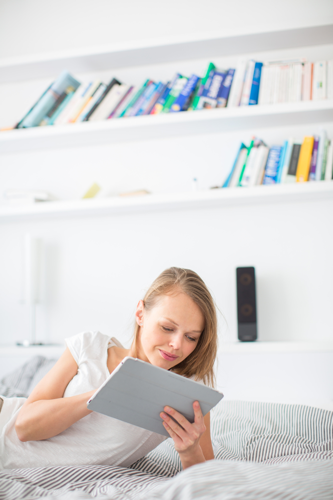 Pretty, young woman lying in bed, using her tablet computer, reading sending e-mails, using apps, while listening to an active bookshelf speaker and having fun